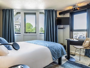 A hotel room with a bed, a desk, and a chair. There are windows with curtains, a view of buildings outside, and a cityscape visible through the windows. - Ibis Styles Boulogne Centre Cathédrale (Boulogne-sur-Mer)