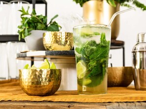 Un bicchiere di bevanda fredda con lime e menta, una ciotola dorata e uno shaker su un piano di legno.