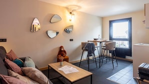 A modern living room with a sofa, a coffee table, a dining area with chairs, a small table with a lamp, and a large window. - Ibis Styles Boulogne Centre Cathédrale (Boulogne-sur-Mer)