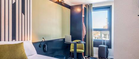 Una habitación de hotel con una cama, un escritorio con una computadora portátil, una silla, una maleta y vistas a edificios a través de una ventana.