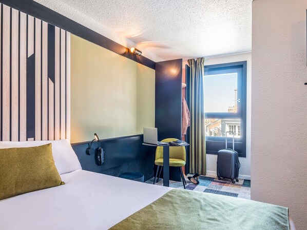 Una habitación de hotel con una cama, un escritorio con una computadora portátil, una silla, una maleta y vistas a edificios a través de una ventana.