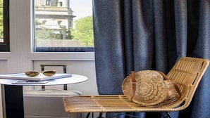 A room with a view of a classical building, a wicker chair, a small table with sunglasses and a notebook, and a window with dark curtains. - Ibis Styles Boulogne Centre Cathédrale (Boulogne-sur-Mer)