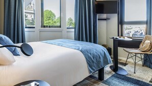 A hotel room with a bed, a desk, and a chair. There are windows with curtains, a view of buildings outside, and a ceiling with recessed lighting. - Ibis Styles Boulogne Centre Cathédrale (Boulogne-sur-Mer)