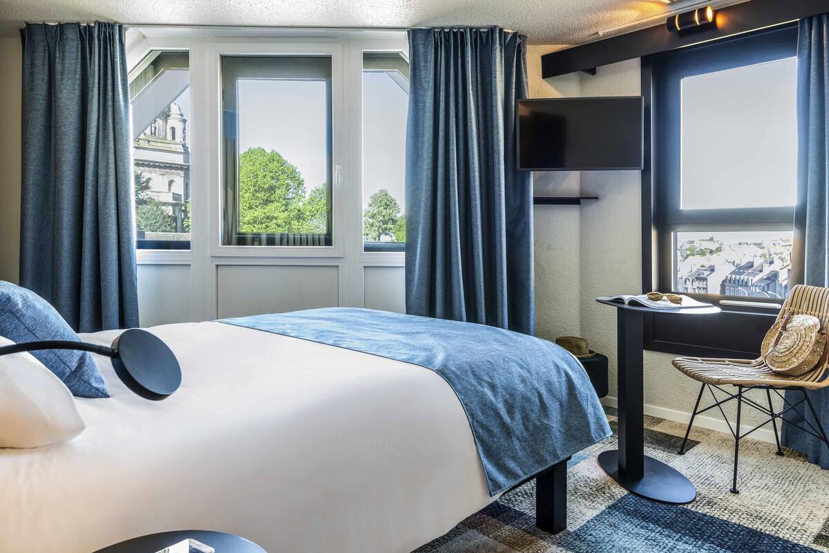 Una camera d'albergo con un letto, una scrivania e una sedia. Sono presenti finestre con tende, vista su edifici e un soffitto con illuminazione a incasso.