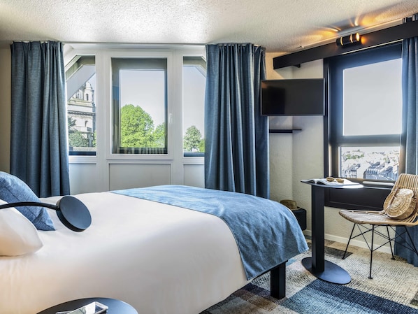 A hotel room with a bed, a desk, and a chair. There are windows with curtains, a view of buildings outside, and a ceiling with recessed lighting.