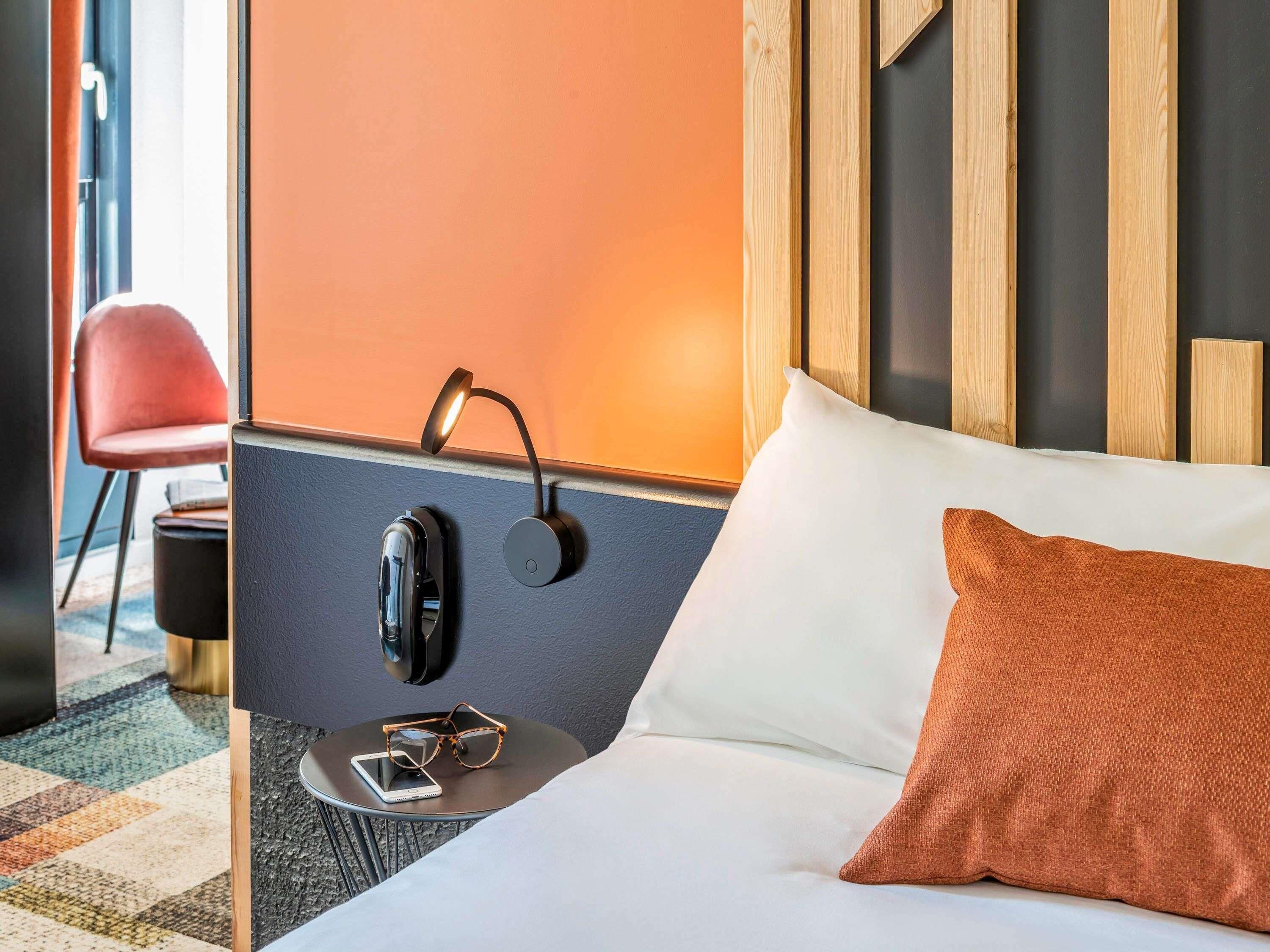 Habitación de hotel con cama, mesita de noche, lámpara de pared, una silla y una pequeña mesa con un teléfono inteligente y gafas.