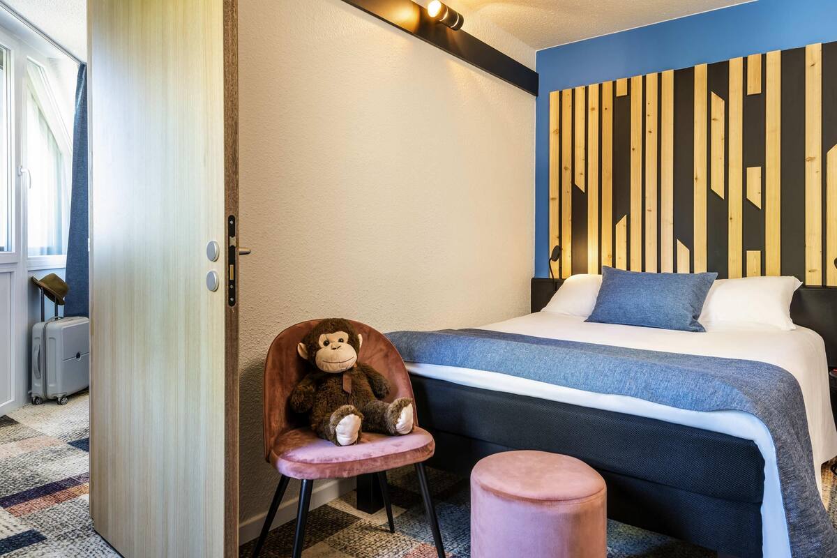Una camera da letto con un letto, una sedia con un peluche, un comodino e una parete con listelli di legno verticali.