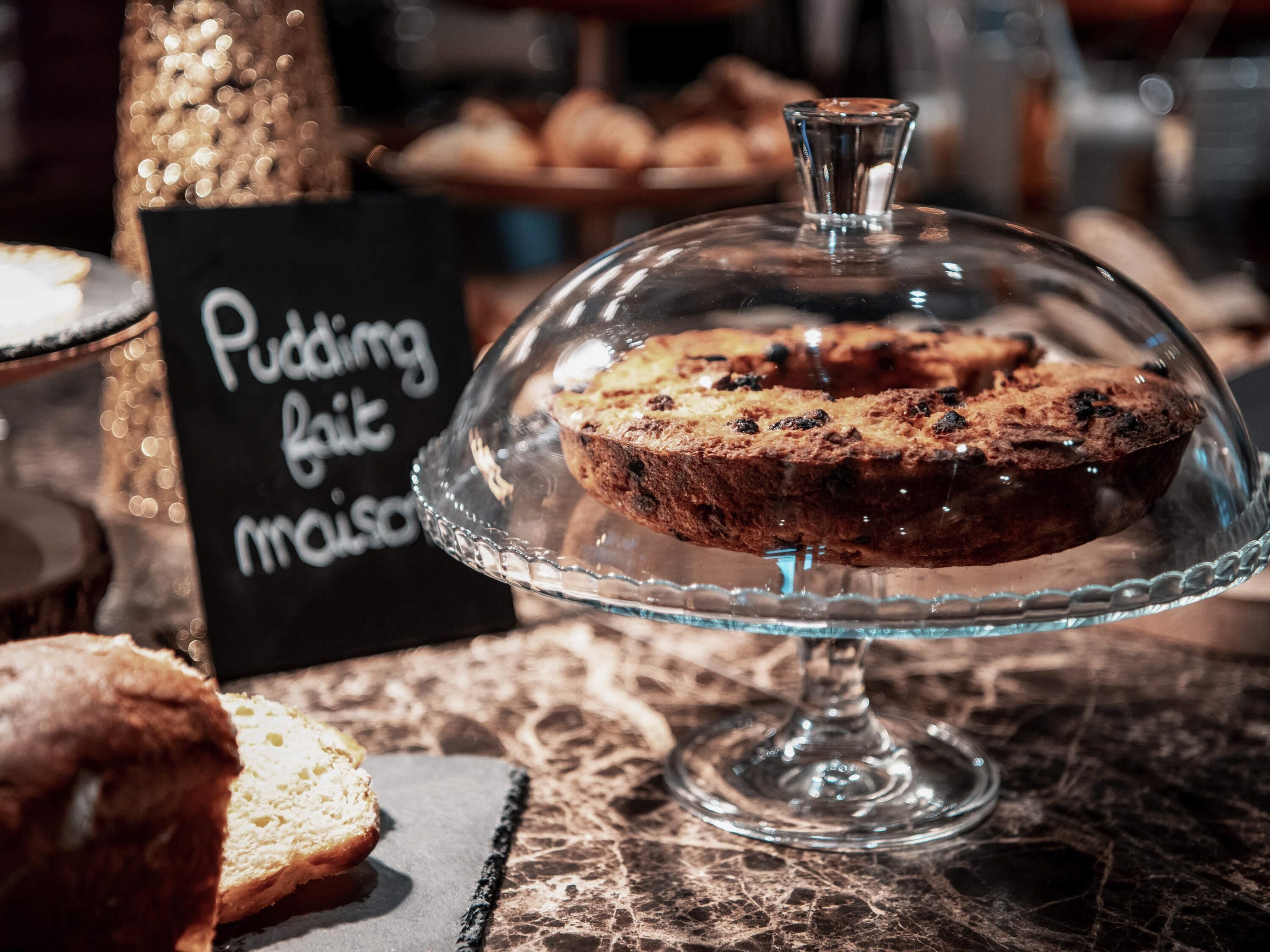 A dessert display with a glass dome cover, a sign reading 'Pudding cake mousse,' and a marble table surface.