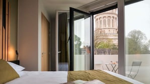 A hotel room with a bed, a magazine on the bed, a window with a view of a building, and a balcony with a table and chairs. - Ibis Styles Boulogne Centre Cathédrale (Boulogne-sur-Mer)