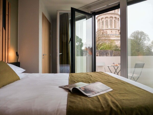 Una camera d'hotel con un letto, una rivista sul letto, una finestra che si affaccia su un edificio e un balcone con tavolo e sedie.