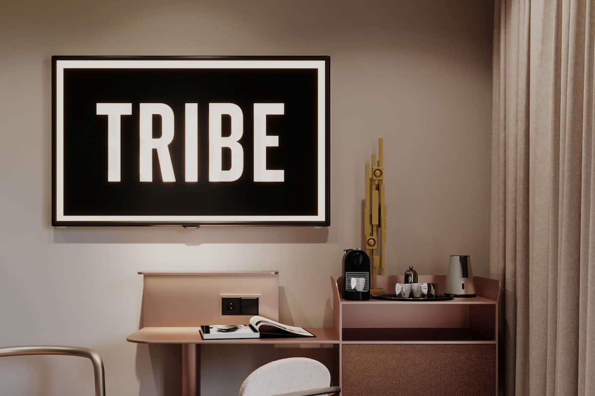A modern room with a framed 'TRIBE' sign, a desk with a chair, a coffee maker, and a shelf with cups.