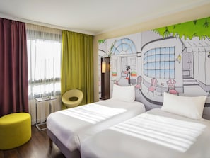 A hotel room with two beds, a yellow ottoman, a chair, and a mural on the wall. - ibis Styles Evry Courcouronnes Hotel and Events (Évry-Courcouronnes)