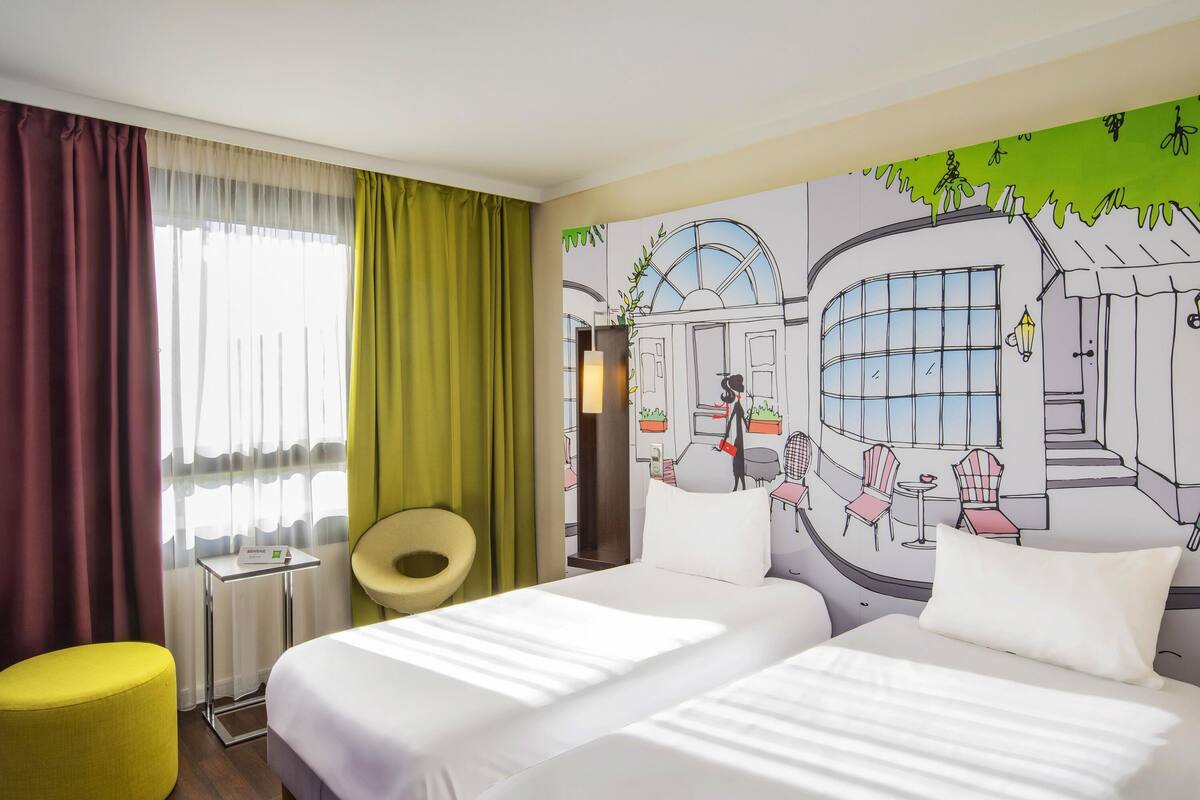 A hotel room with two beds, a small table, a chair, and a large window with curtains.