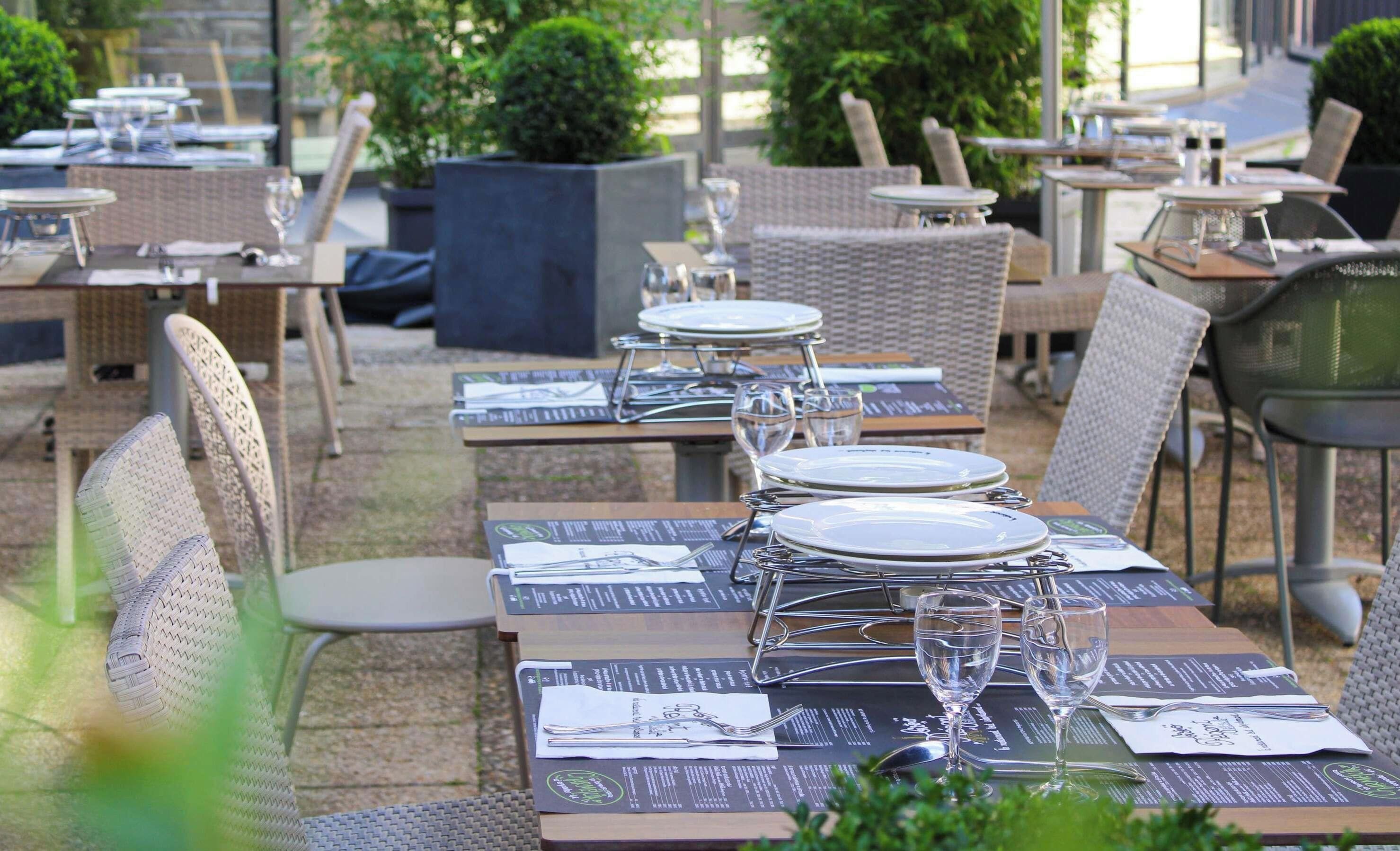 An outdoor dining area with tables set for two, each with a menu, cutlery, and wine glasses.