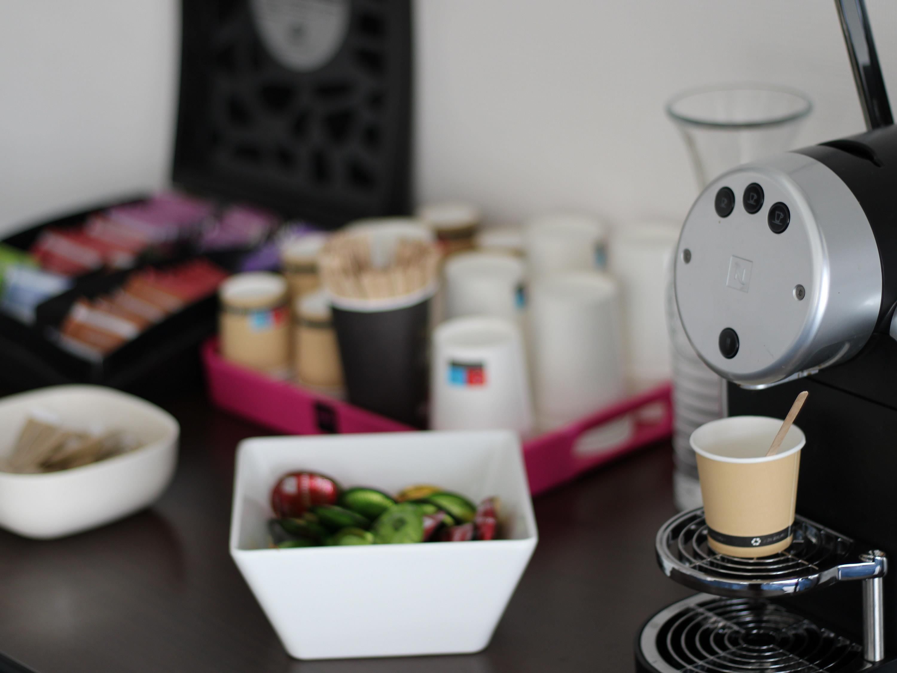Een koffieapparaat met kopjeshouder, een schaal met fruit en een bak koffiecups op een tafel.