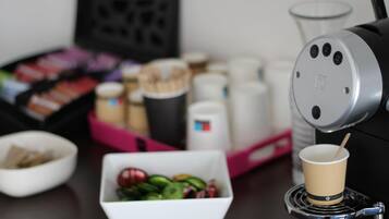 Een koffieapparaat met kopjeshouder, een schaal met fruit en een bak koffiecups op een tafel.