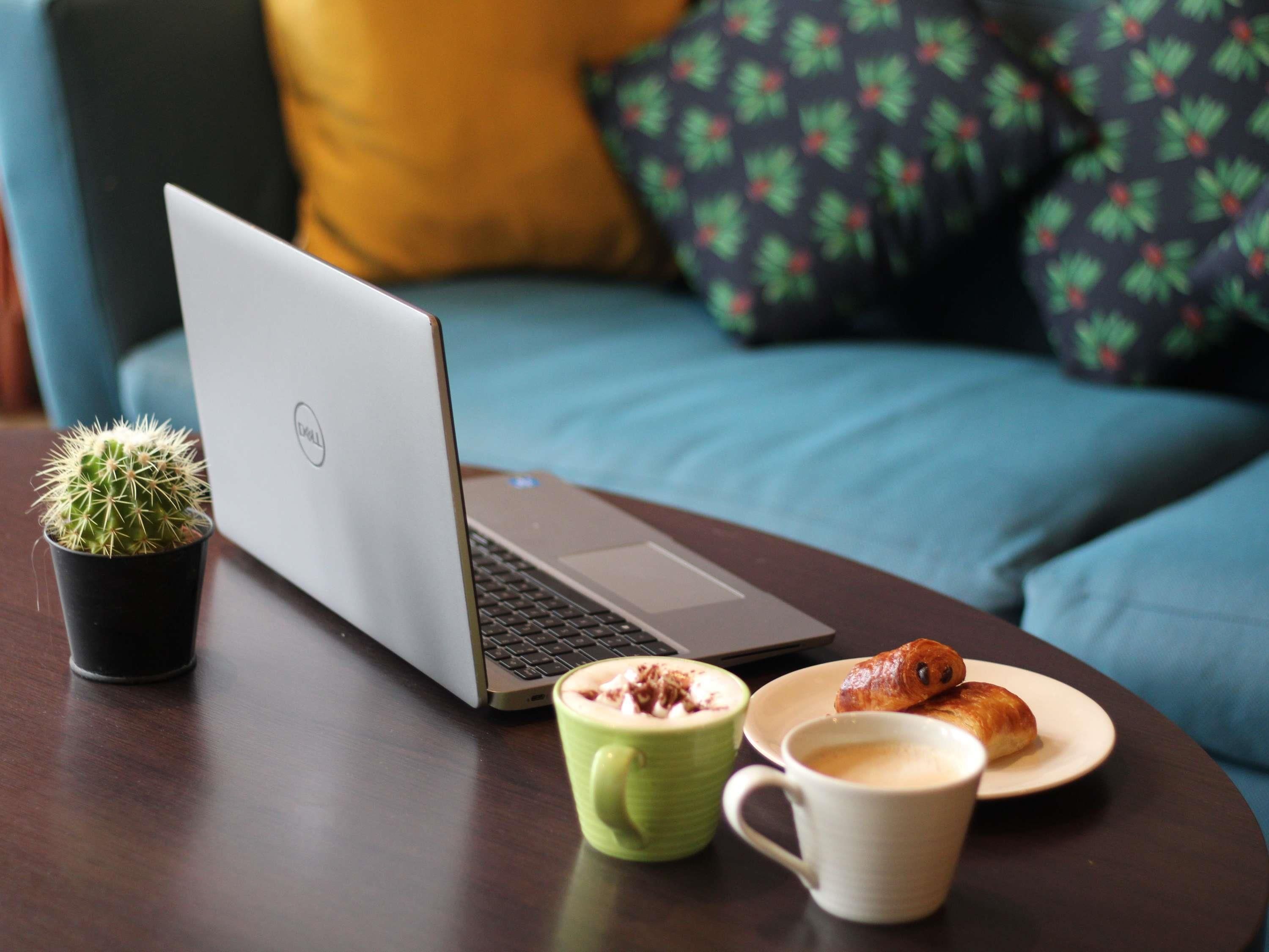 Een laptop op een tafel met een cactus, een groene mok, een witte mok en een bord met gebak.