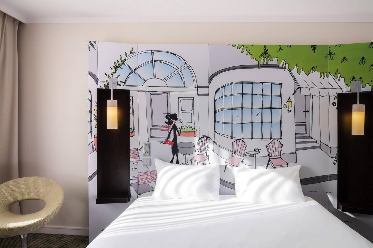 Ein Hotelzimmer mit einem Bett, einem Sessel und einer großen Wandteppich-Szene, die einen stilisierten Innenraum zeigt.