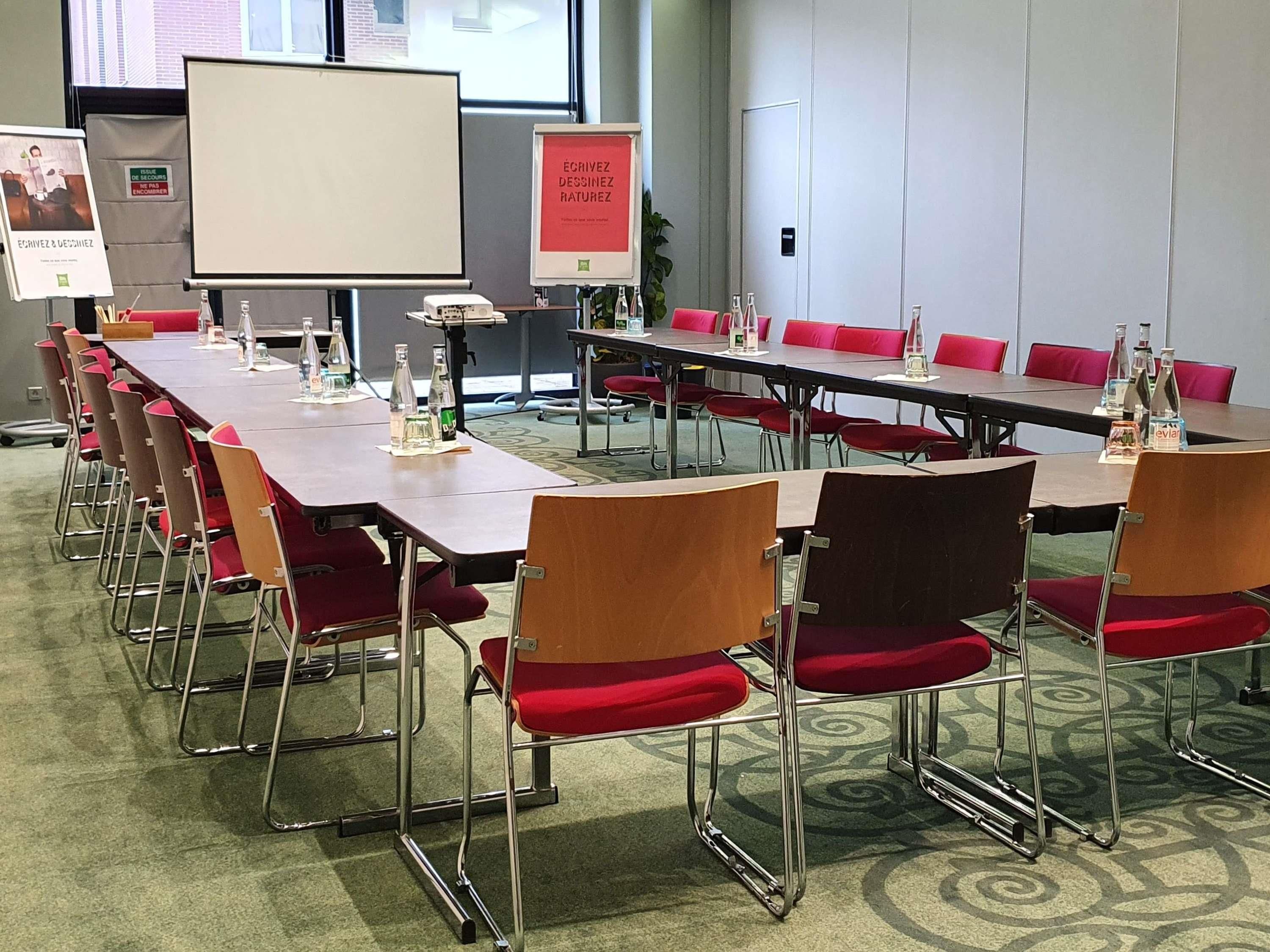 A conference room with long tables, red chairs, a whiteboard, and a projector screen.