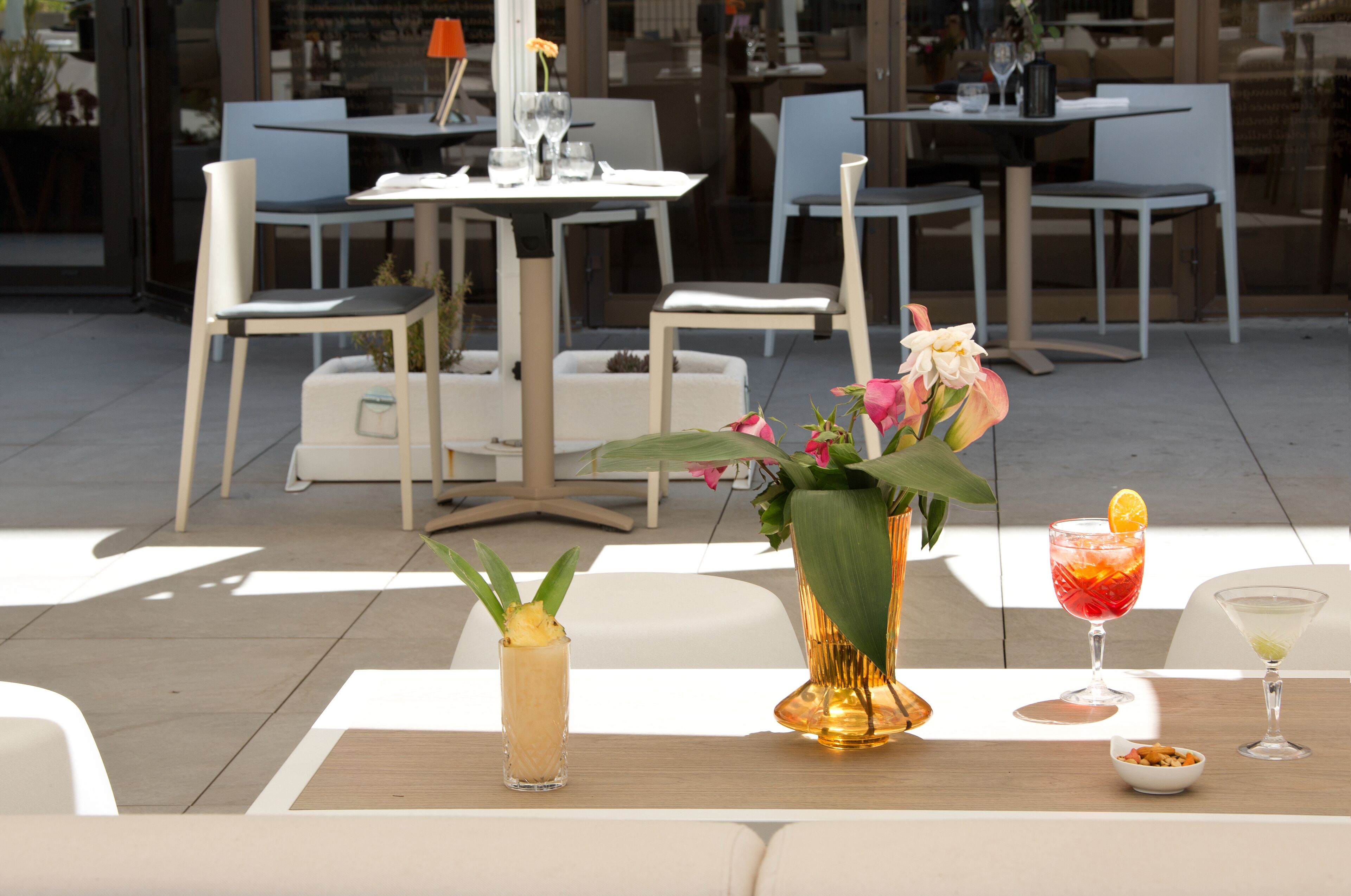 An outdoor dining area with tables and chairs, a vase with flowers, and two drinks on the table.