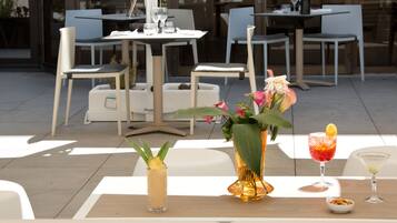 An outdoor dining area with tables and chairs, a vase with flowers, and two drinks on the table.