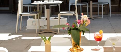 An outdoor dining area with tables and chairs, a vase with flowers, and two drinks on the table.