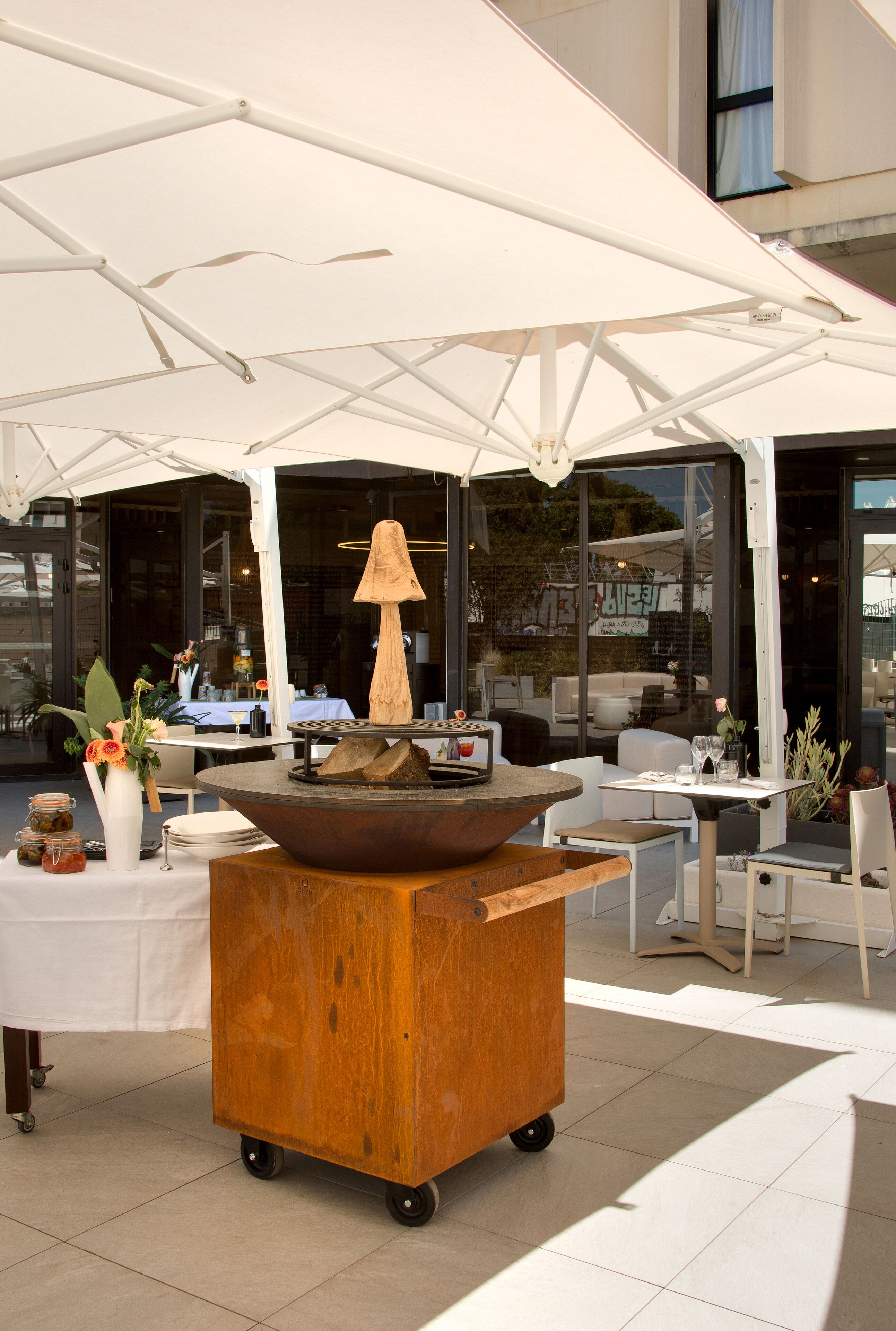 An outdoor seating area with a central fire feature, tables, chairs, and a white umbrella structure.