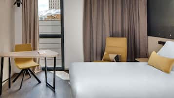 Une chambre d’hôtel avec un lit, un bureau, une chaise et une vue sur un immeuble par la fenêtre.