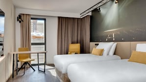 Une chambre d’hôtel avec deux lits, un bureau, une chaise et une grande fenêtre donnant sur des immeubles.