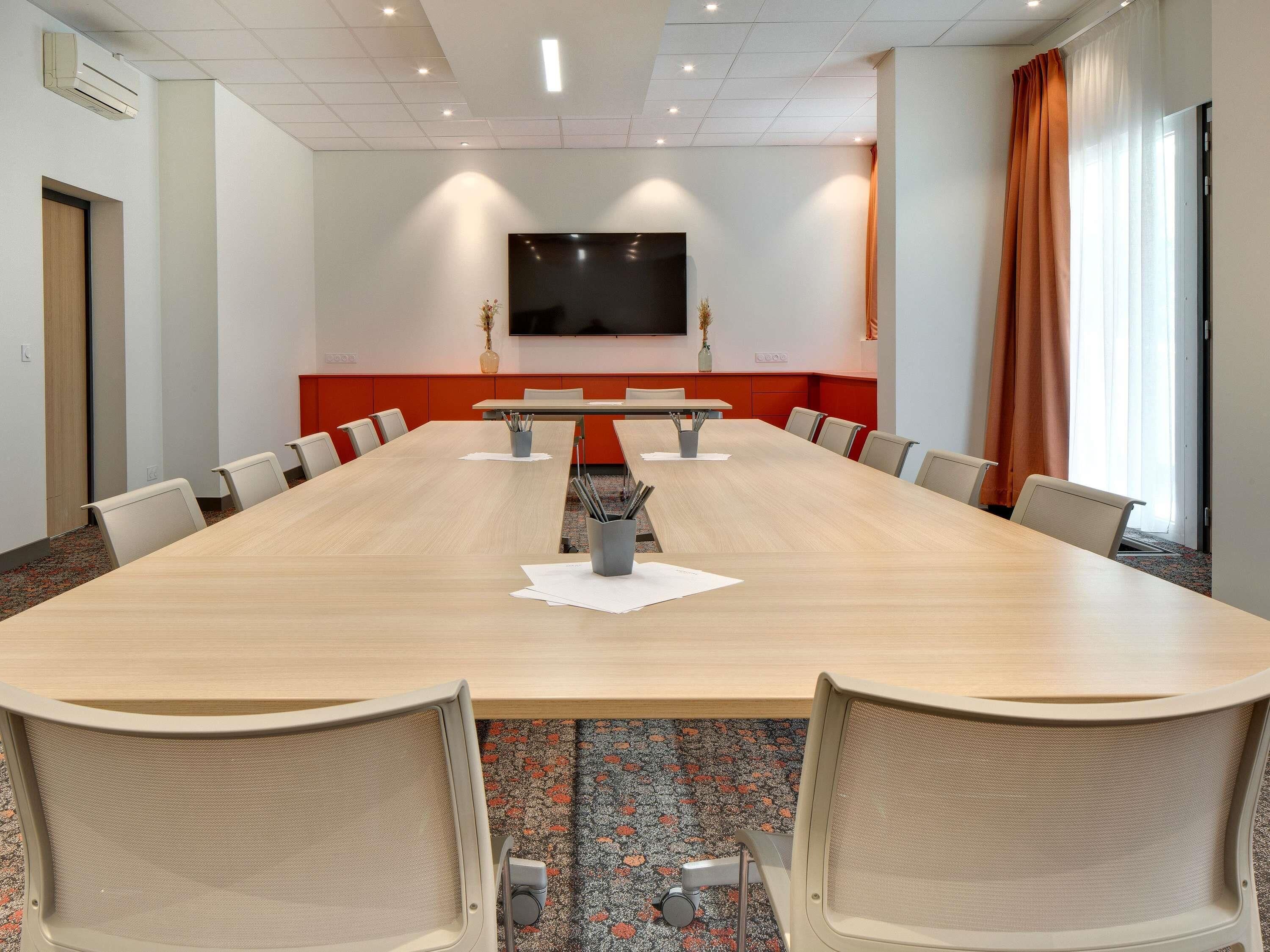 A conference room with a long table, chairs, a flat-screen TV, and a red accent wall.