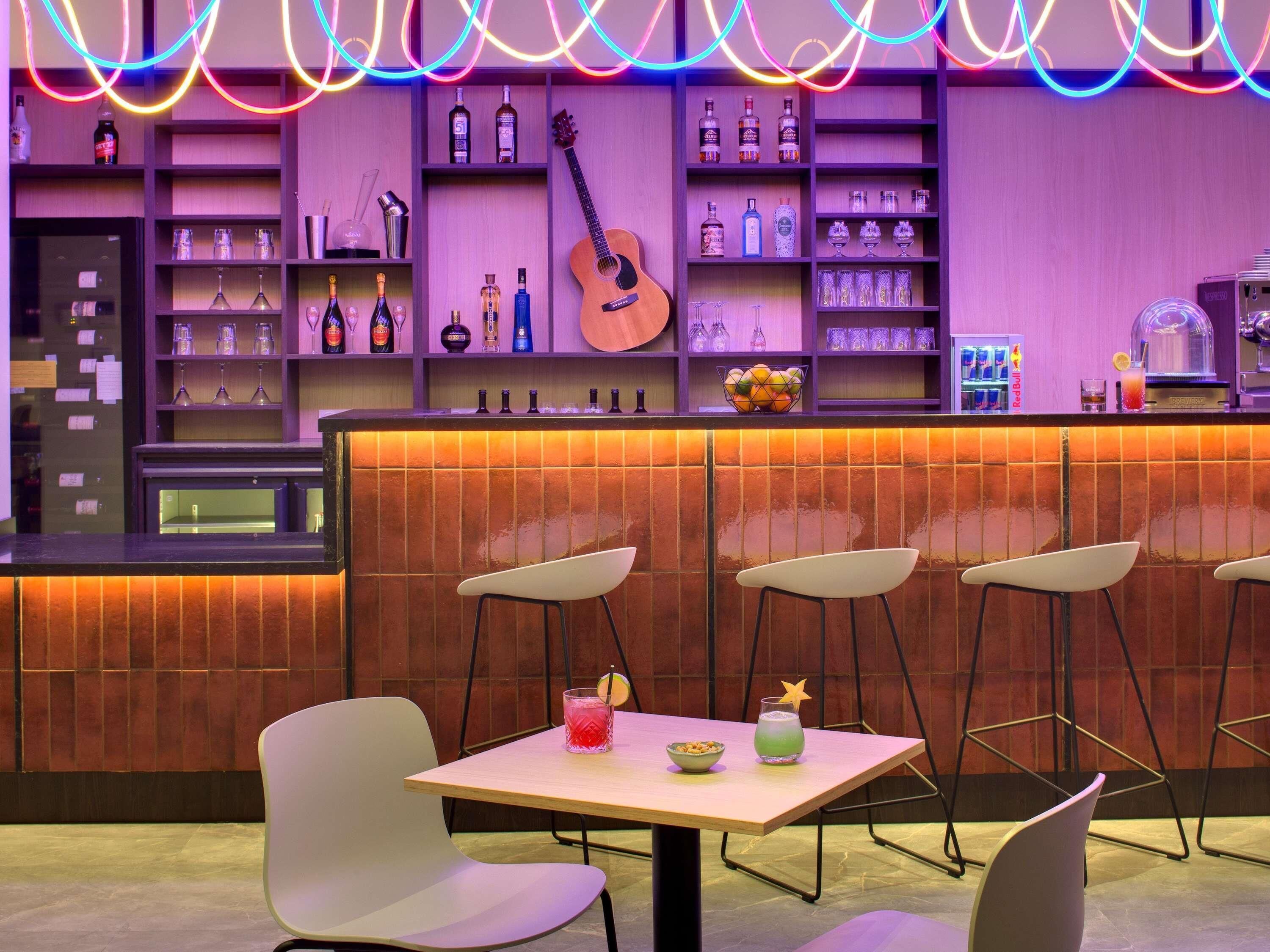 A bar area with a wooden counter, shelves with bottles, glasses, and a guitar. There are four white chairs around a table with drinks.