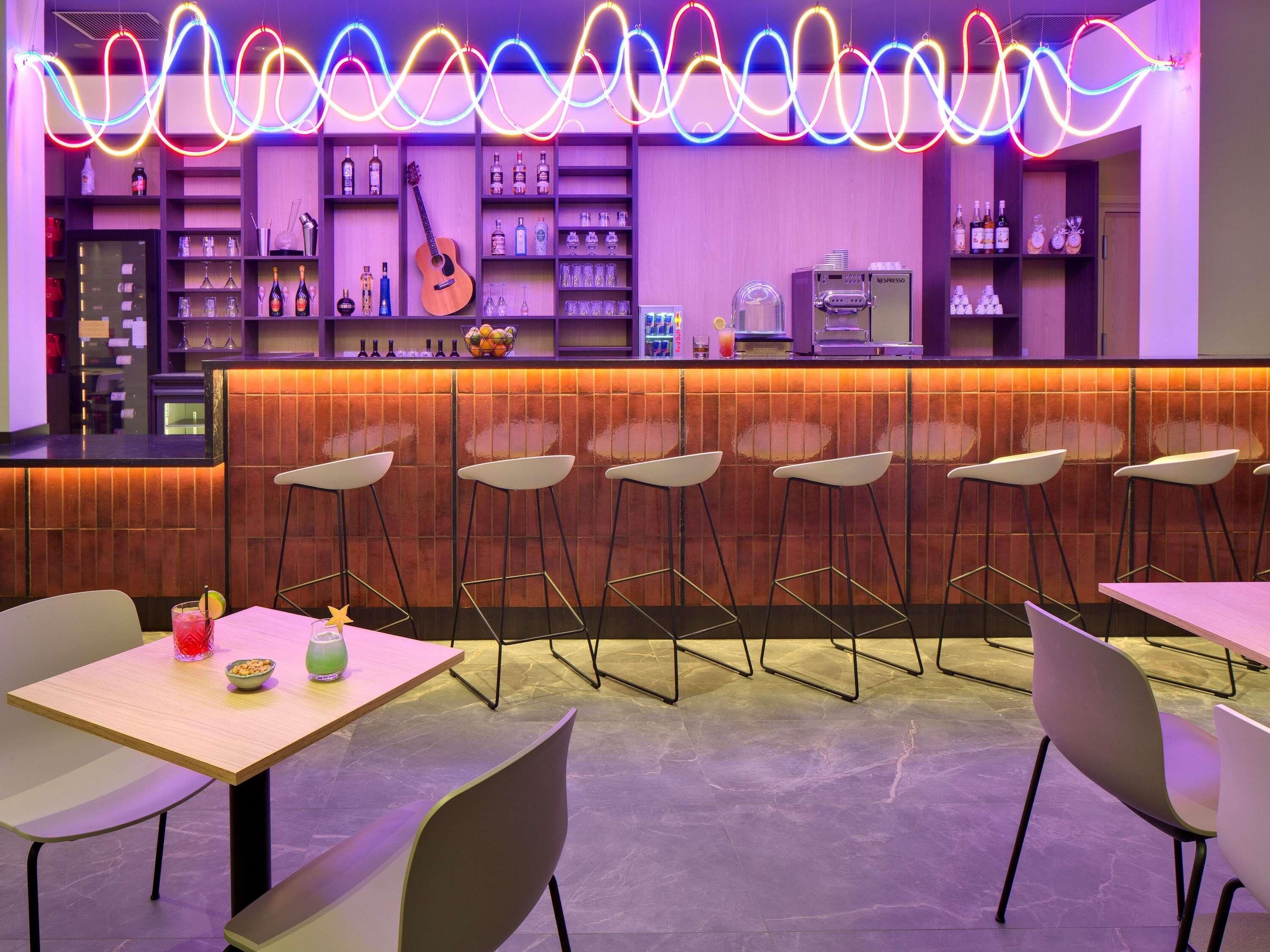 A bar area with neon lights, a guitar, shelves with bottles, and bar stools.