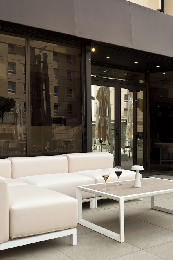 Un área de estar al aire libre con un sofá blanco y una mesa, una puerta de vidrio que refleja el edificio de fondo, y una lámpara blanca sobre la mesa.