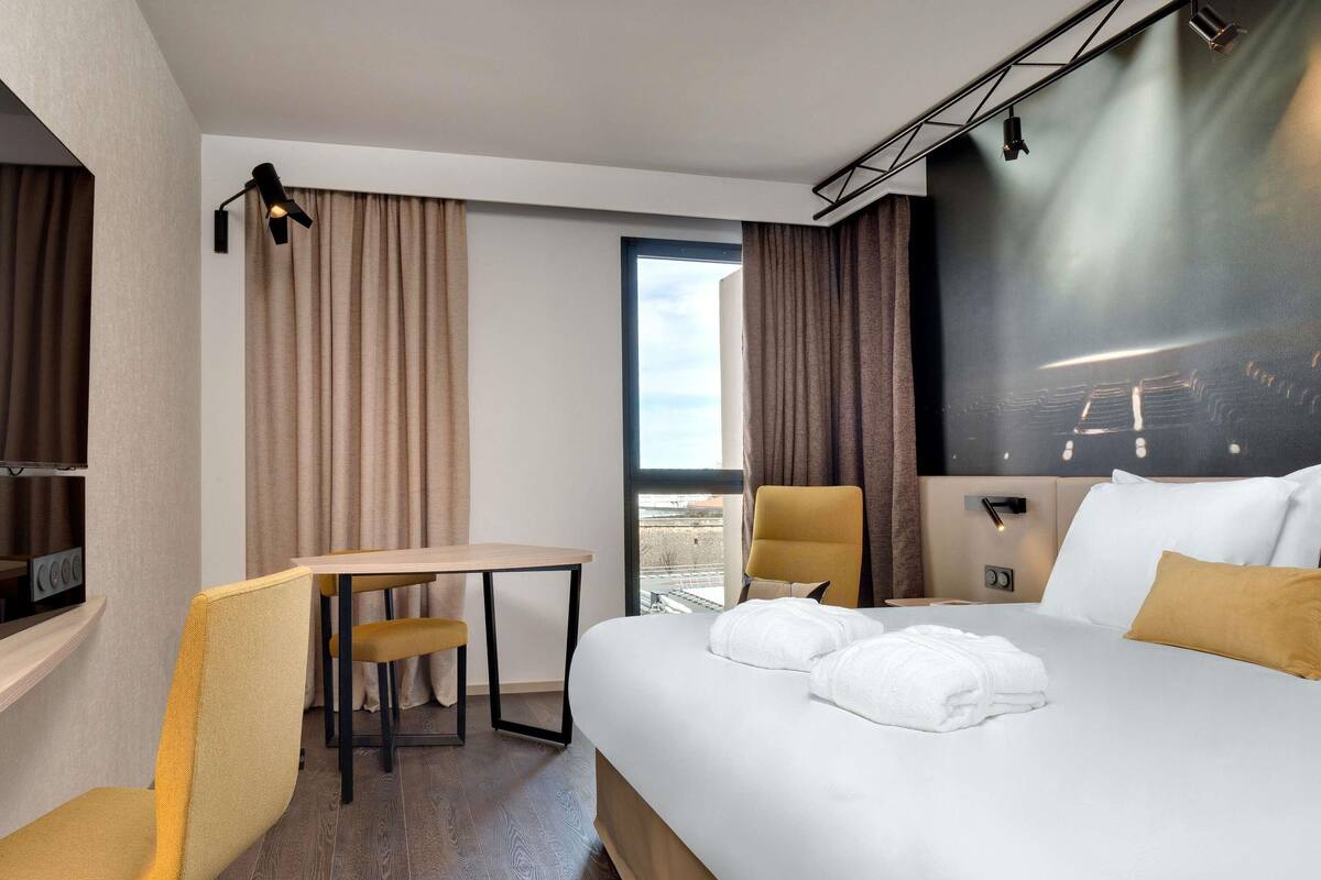 Habitación de hotel con cama, escritorio, silla, televisor y ventana grande.