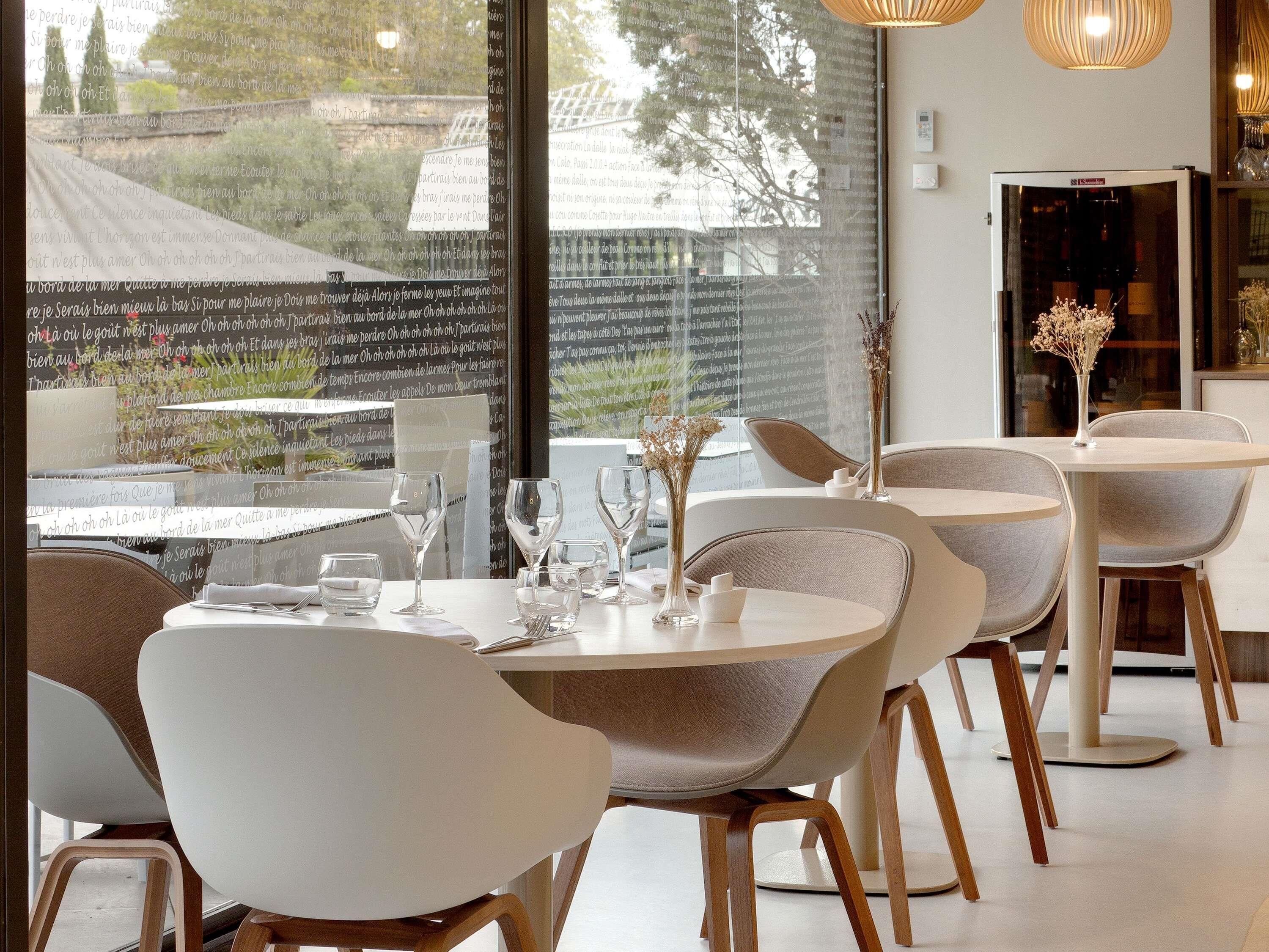 A modern dining area with round tables and chairs, a view of the outside through large windows, and a wine cooler in the background.