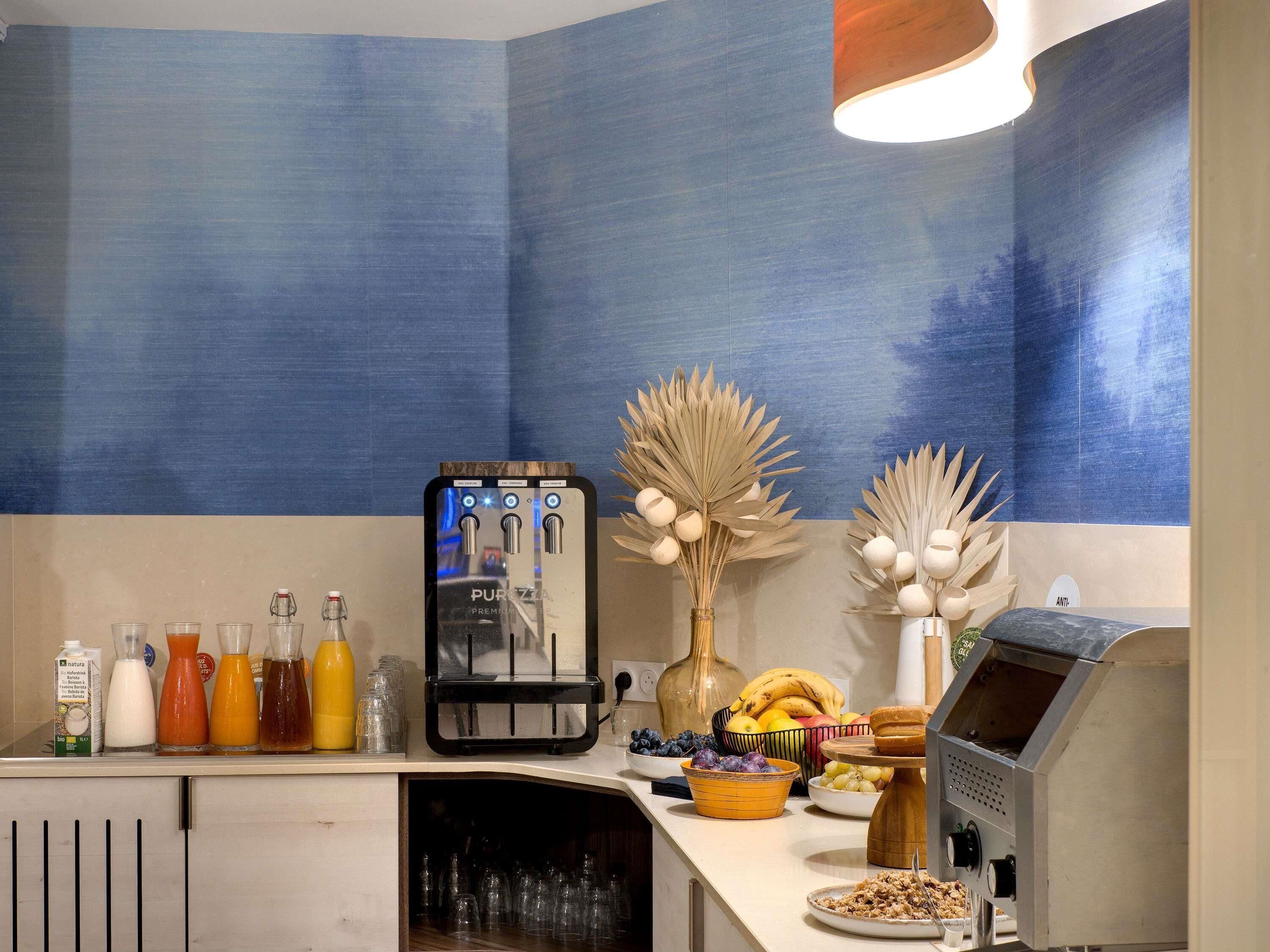 A kitchen counter with a coffee machine, various drink bottles, a fruit bowl, and a decorative vase.