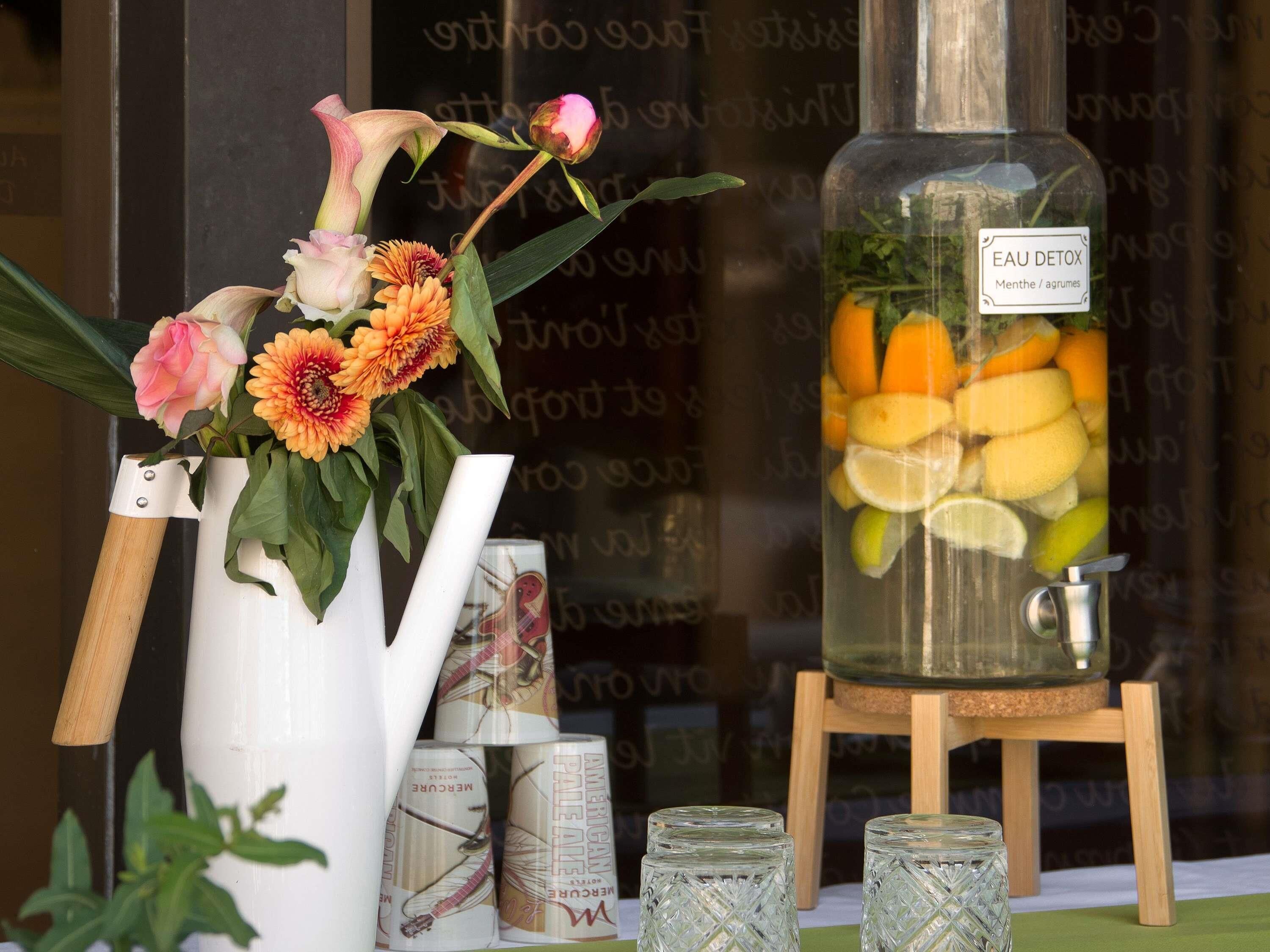 Une composition décorative comprenant des fleurs, un récipient en verre rempli de fruits et une bouteille étiquetée « EAU DÉTOX ».