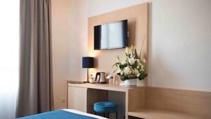 Habitación de hotel con una cama, un escritorio con televisor de pantalla plana, una lámpara, un florero con flores y un cojín azul.