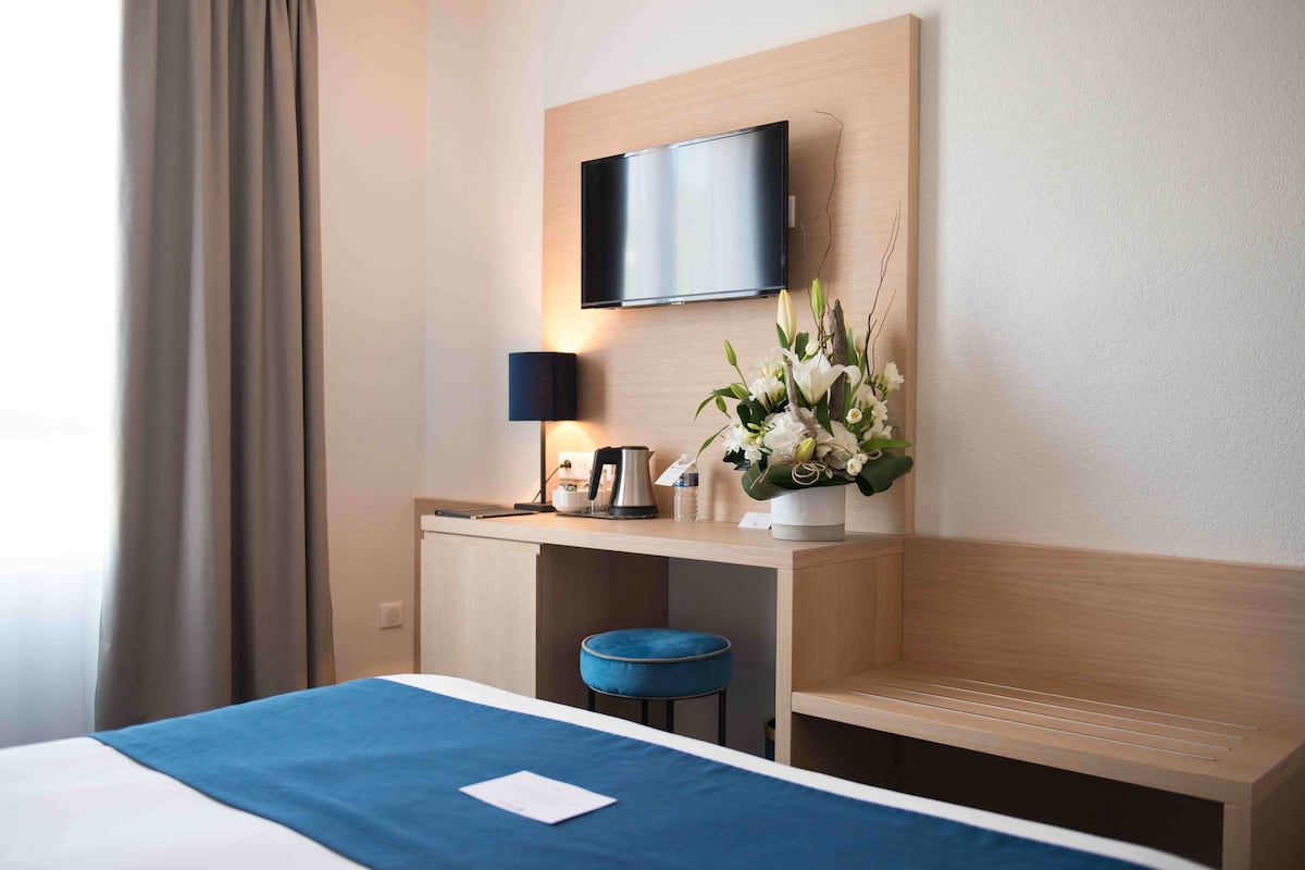 A hotel room with a bed, a desk with a flat-screen TV, a lamp, a vase with flowers, and a blue cushion.