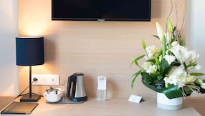 Habitación de hotel con escritorio, televisor de pantalla plana montado en la pared, lámpara, cafetera, botella de agua, arreglo floral y un taburete azul.