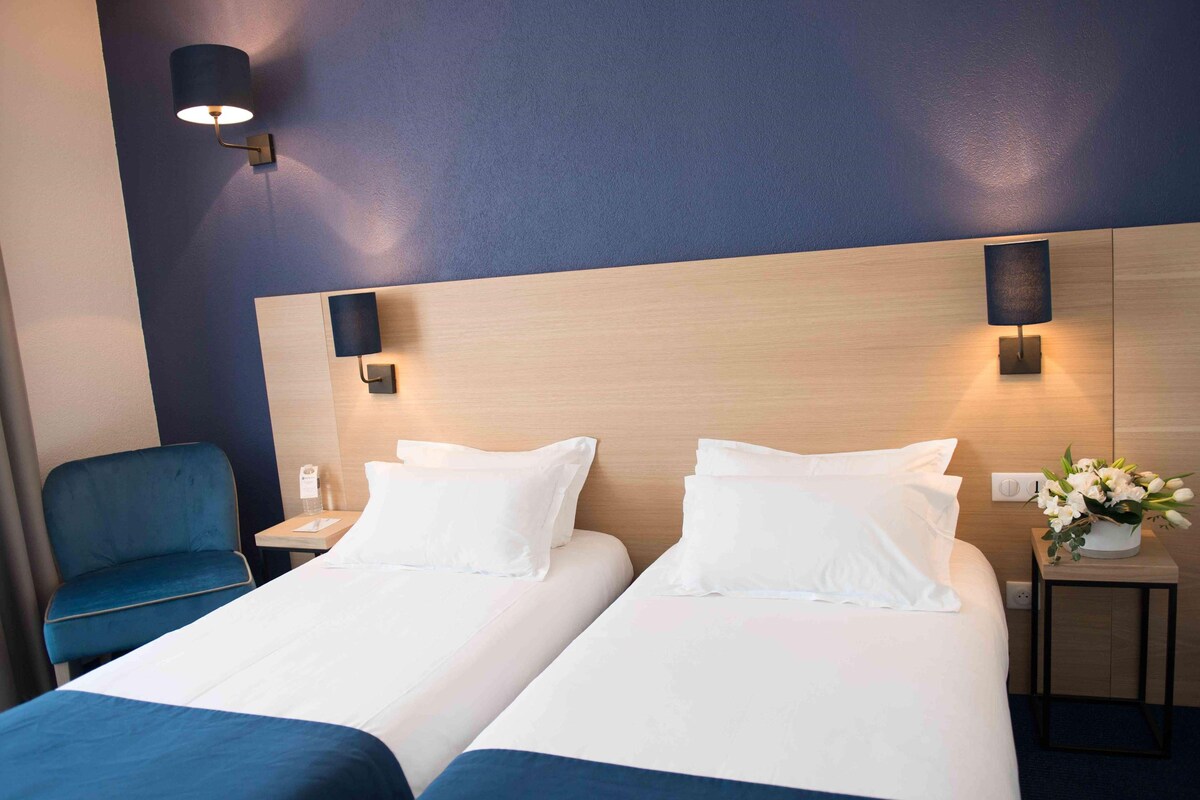 Une chambre d’hôtel avec deux lits, un fauteuil bleu, des tables de chevet avec des lampes et une petite table sur laquelle est posé un vase de fleurs.
