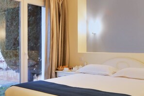 A hotel room with a bed, a nightstand with a lamp, a window with curtains, and a view of trees outside.