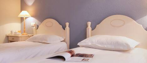 Una cama doble con ropa de cama y almohadas blancas, una mesita de noche con una lámpara, y una revista sobre la cama.