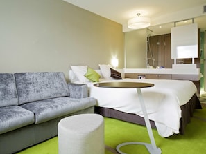 Moderni hotellihuone, jossa on harmaa sohva, pyöreä pöytä, valkoisilla vuodevaatteilla varustettu sänky, vihreä matto ja suuri peili kylpyhuoneen peilipöydän yläpuolella.