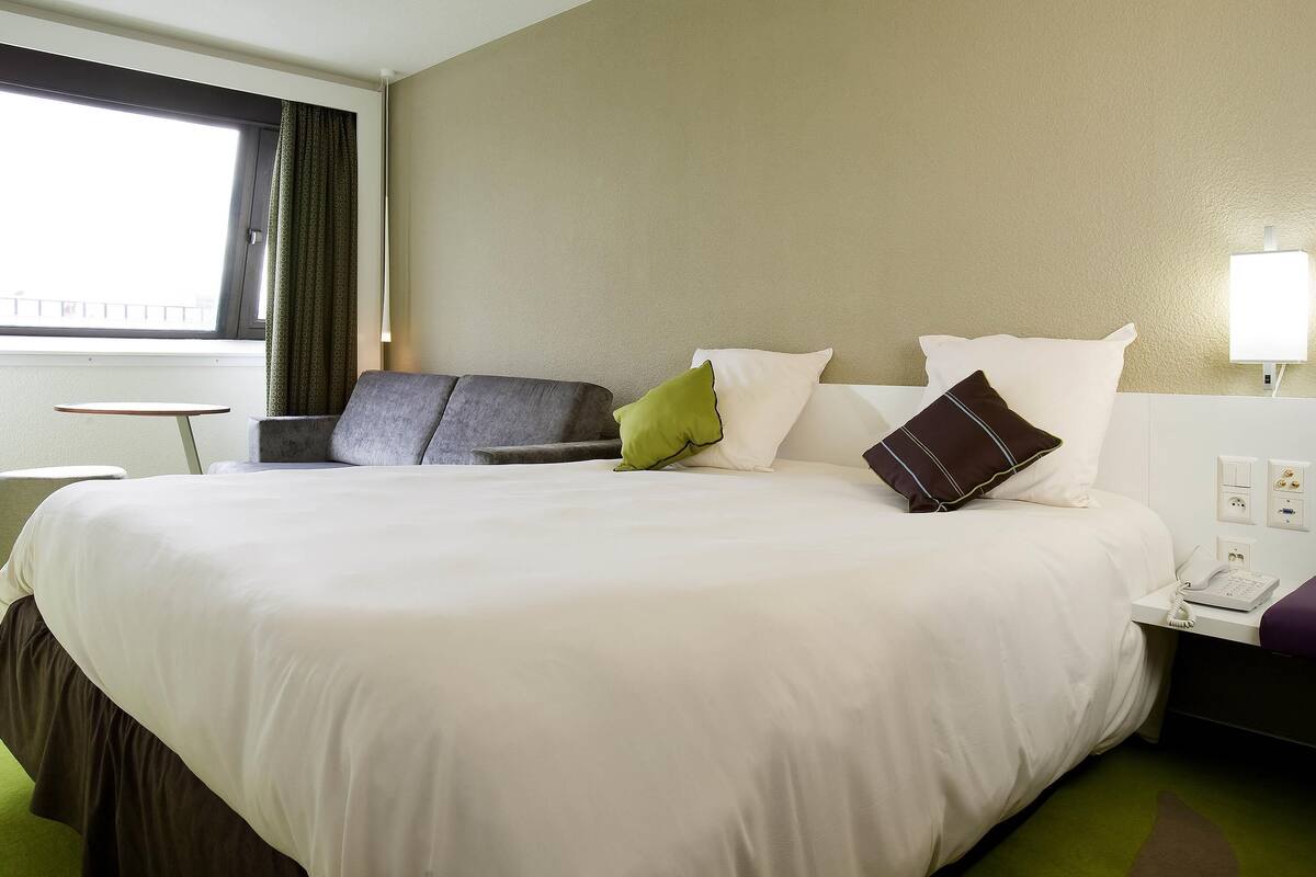 Ein Hotelzimmer mit Bett, einem kleinen Tisch, einem Stuhl, einer Lampe und einem fenster mit Vorhängen.