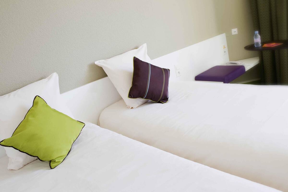 A hotel room with two beds, each with a pillow and a small table with a water bottle.