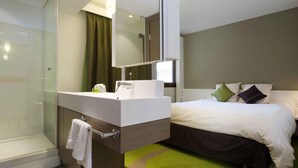 Una habitación de hotel moderna con cabina de ducha de vidrio, tocador blanco con espejo, cama con almohadas verdes y negras, y alfombra verde.