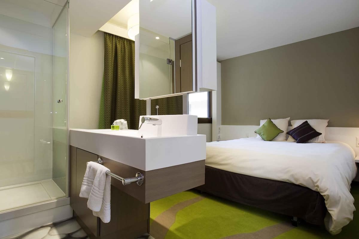 Ein modernes Hotelzimmer mit Duschkabine aus Glas, einem weißen Waschtisch mit Spiegel, einem Bett mit grün-schwarzen Kissen und einem grünen Teppich.
