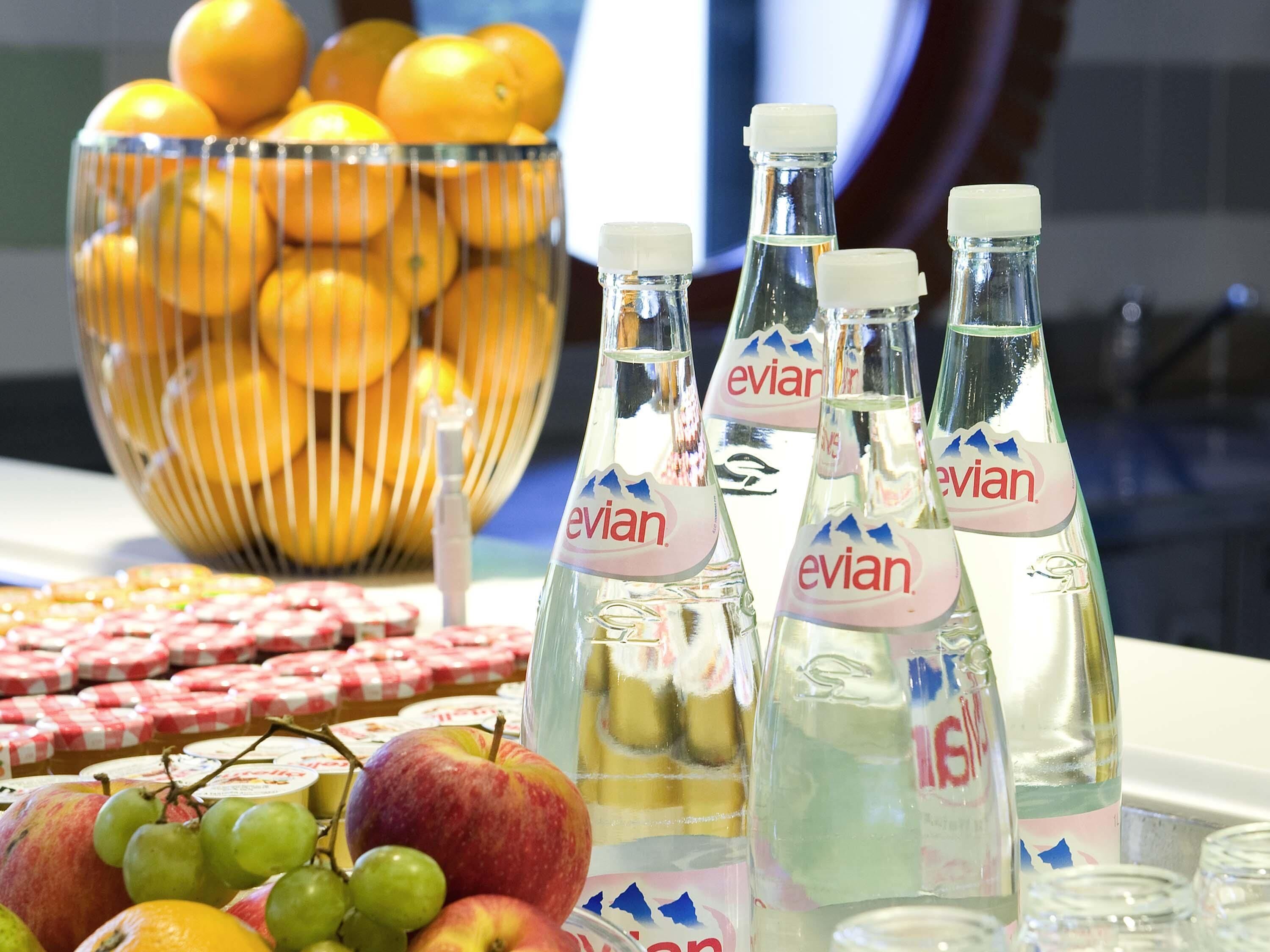 Una mesa con botellas de Evian, un bol de naranjas y diversas frutas y quesos.