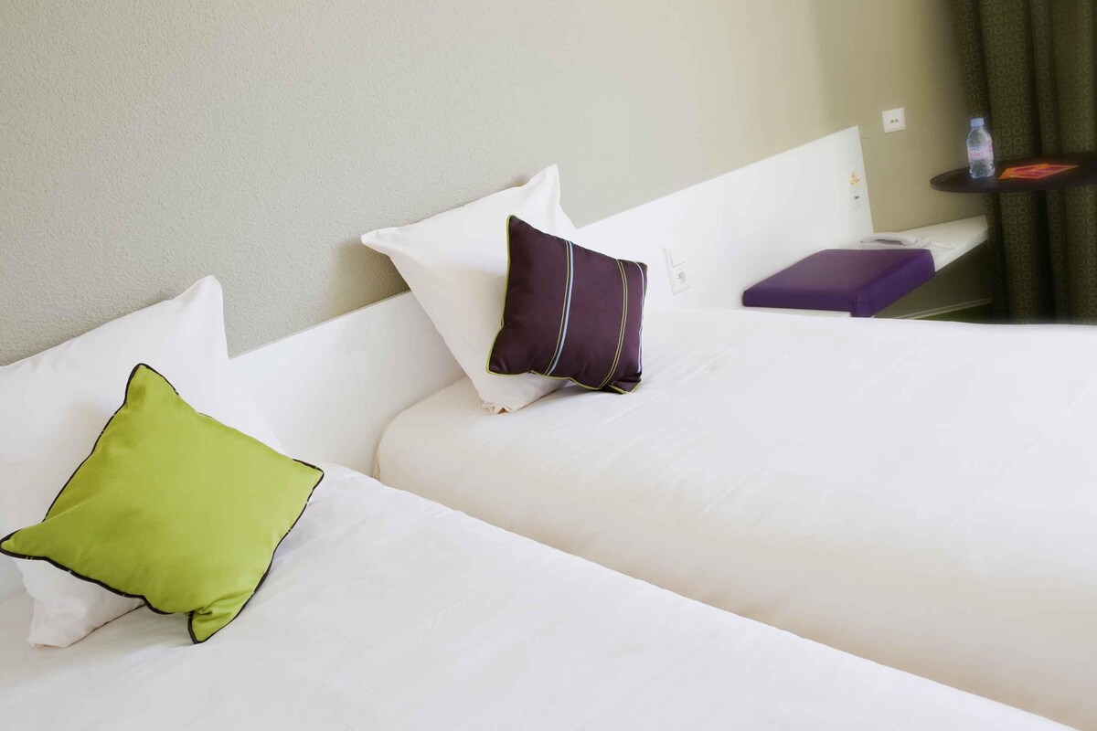 Ein Hotelzimmer mit zwei Betten, jeweils mit einem Kopfkissen und einem kleinen Tisch mit einer Wasserflasche.